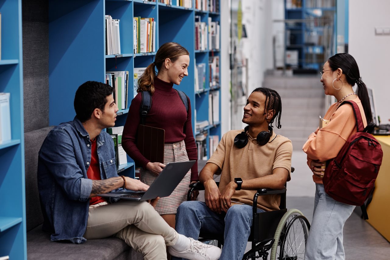 Group of college students with backpacks, books, headphones and a laptop talking in the library.