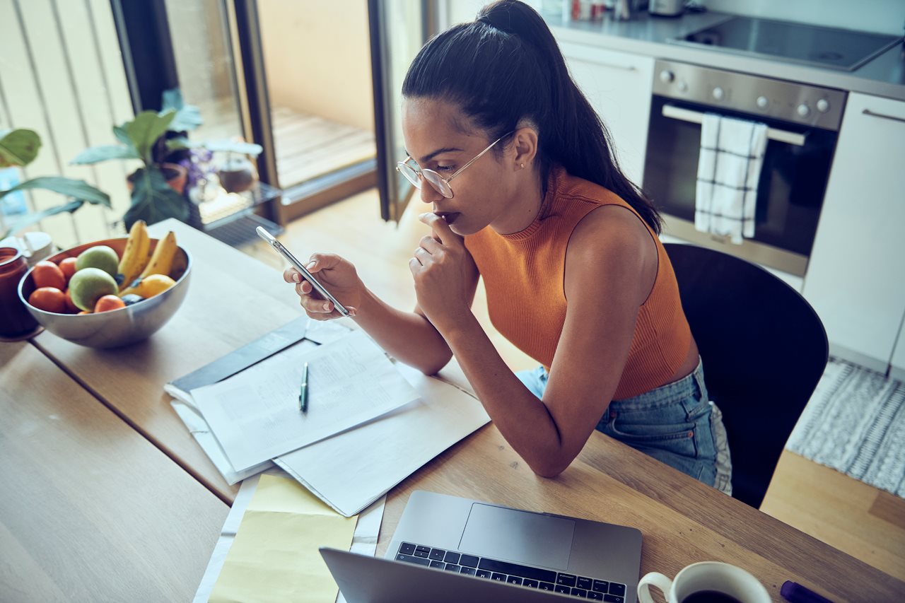 Young woman reviewing paper work with cell phone while sitting at her kitchen table.