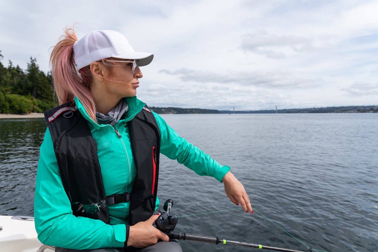 Ashley wearing a teal shirt, life jacket, white hat and sunglasses fishing on a boat in the lake.