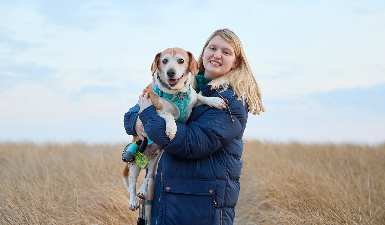 Smiling Tara wearing a coat and hugging her dog while standing in a field.