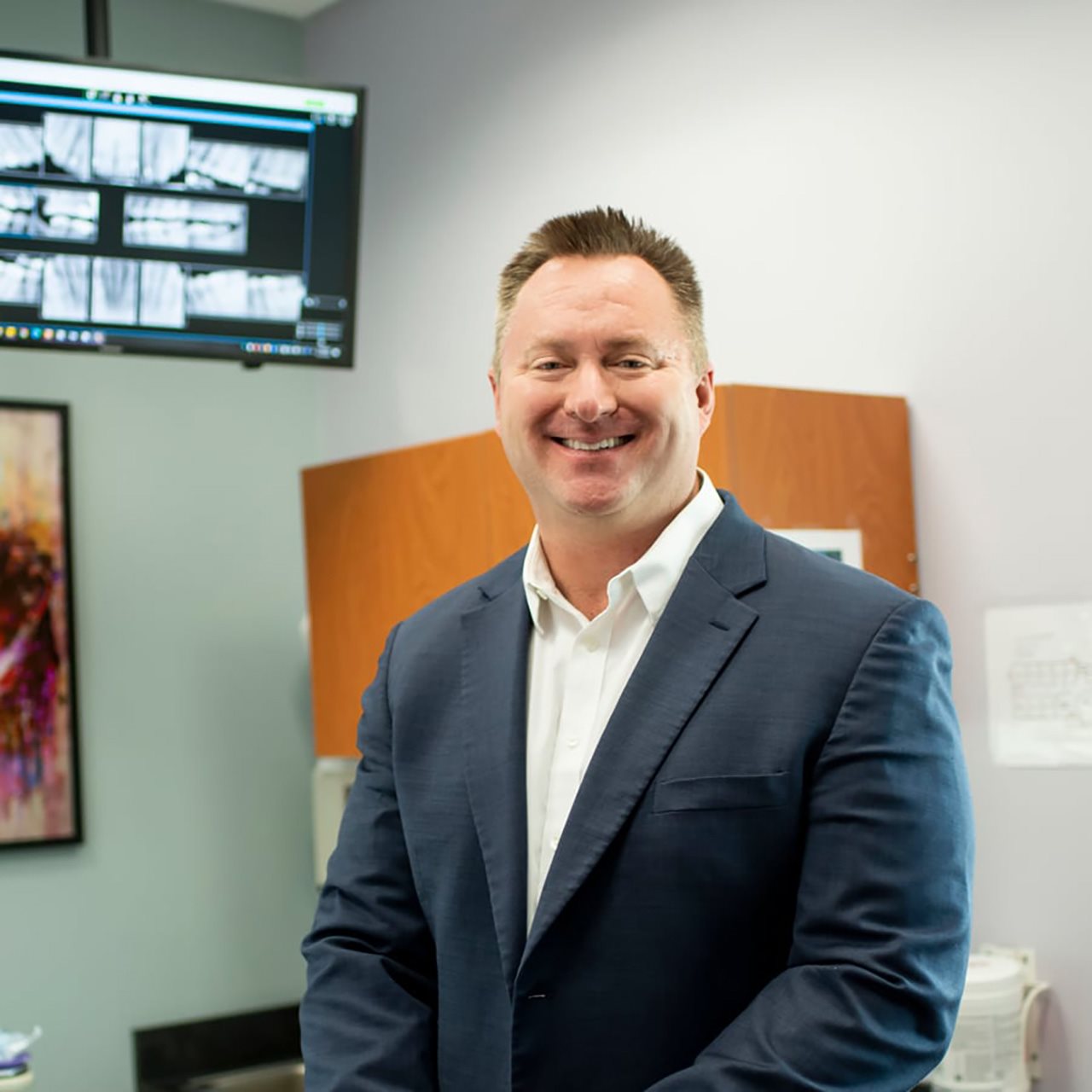 Dr. Peter Drews smiling in an office with x-rays of teeth on a television behind him.