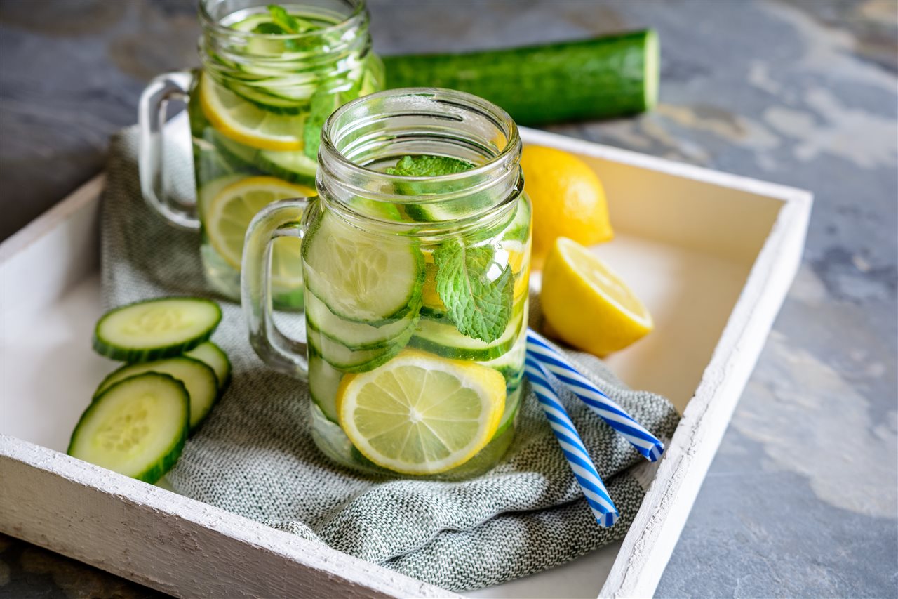 Jelly jar cups with cucumber and lemon water in a tray on a counter top.