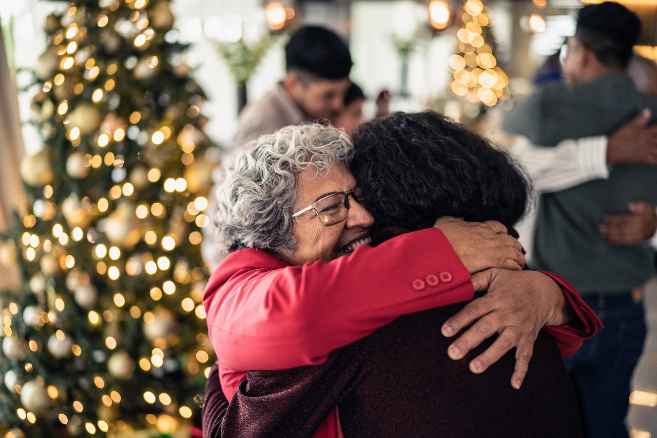 Younger woman hugging her grandmother at a family gathering with other people and a Christmas tree in the background.