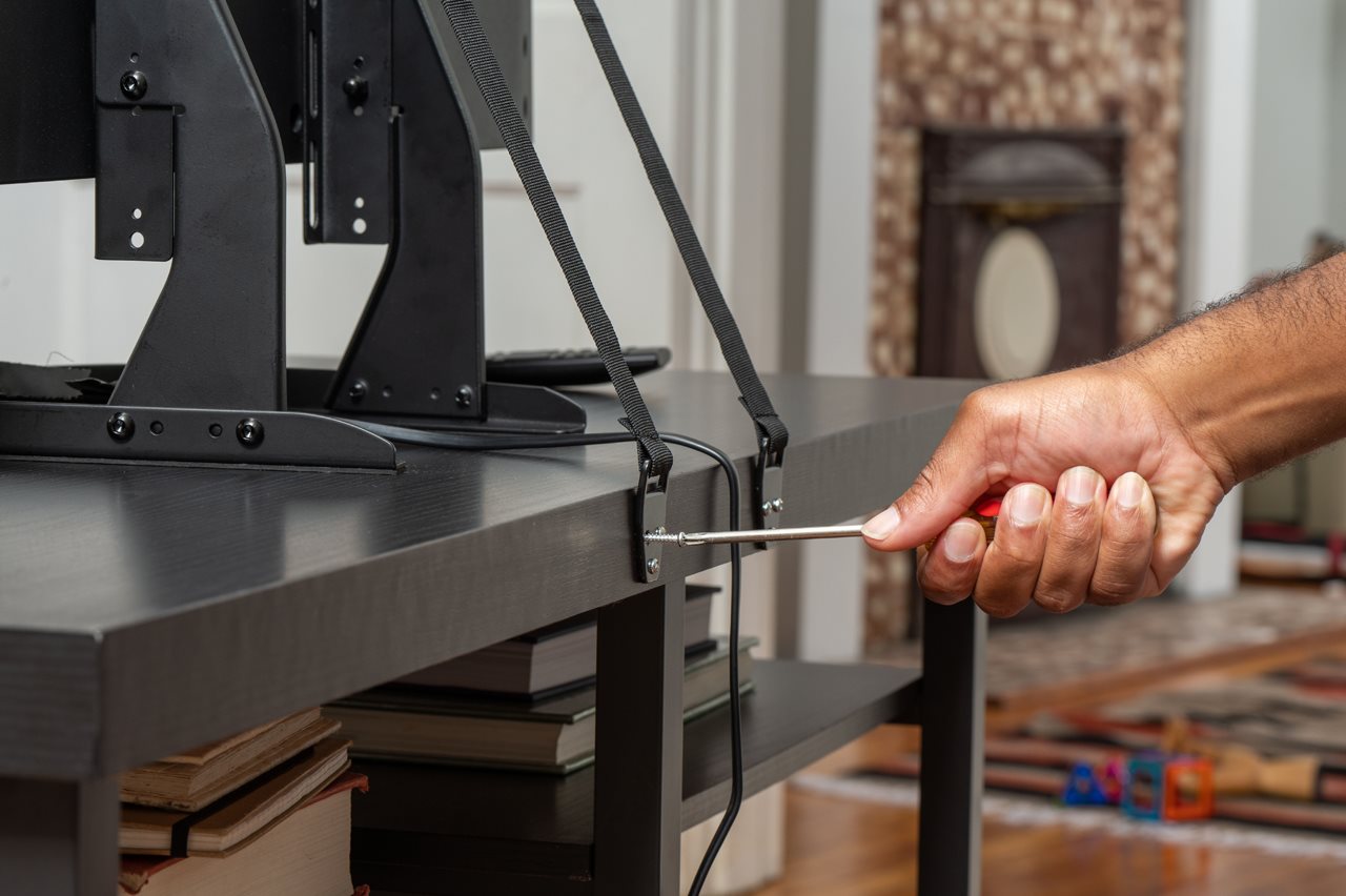 A man securing a flat screen tv to the back of a desk with straps.