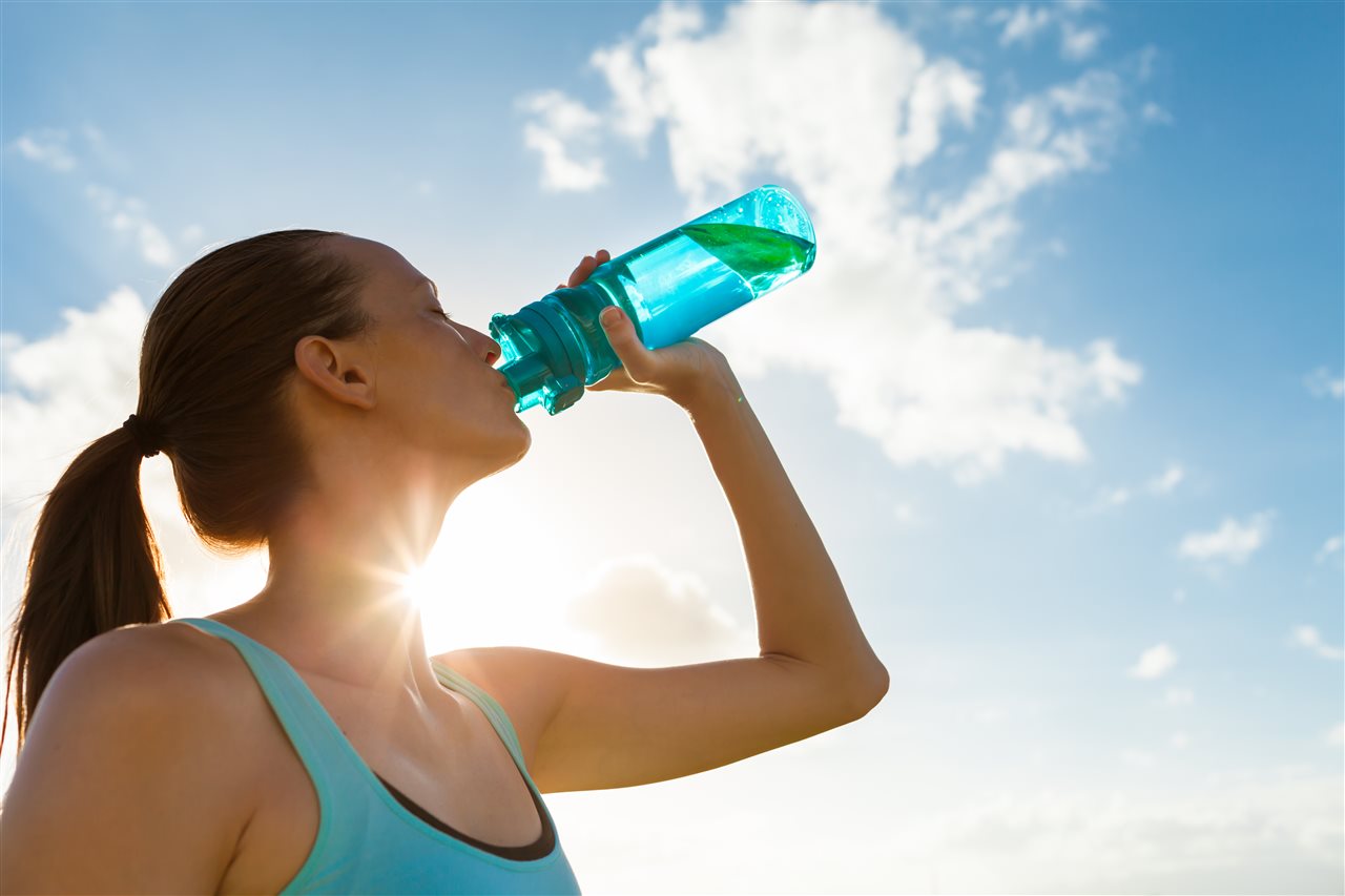 Woman with a pony tail and wearing athletic wear, drinking water from a reuseable bottle.