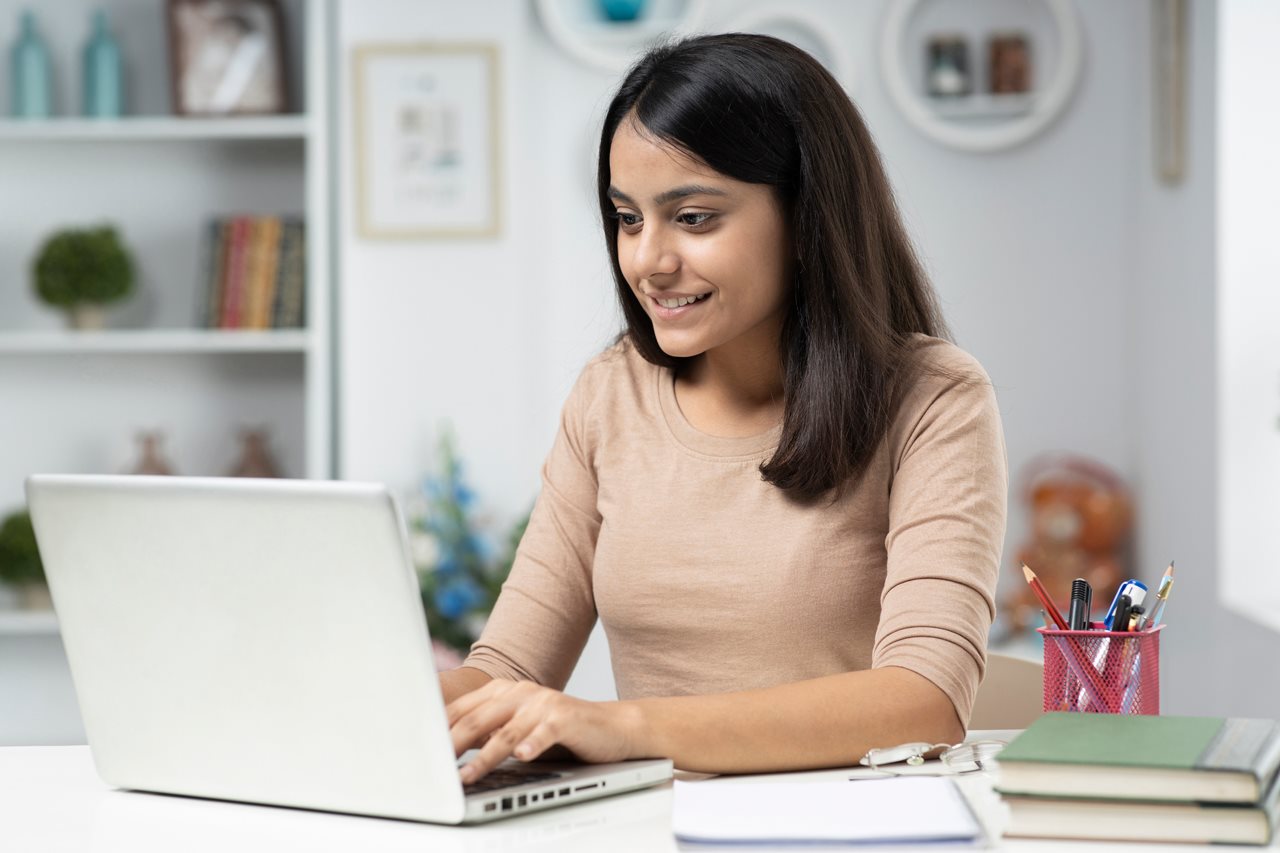 Young woman writing on a laptop at a desk in her room.