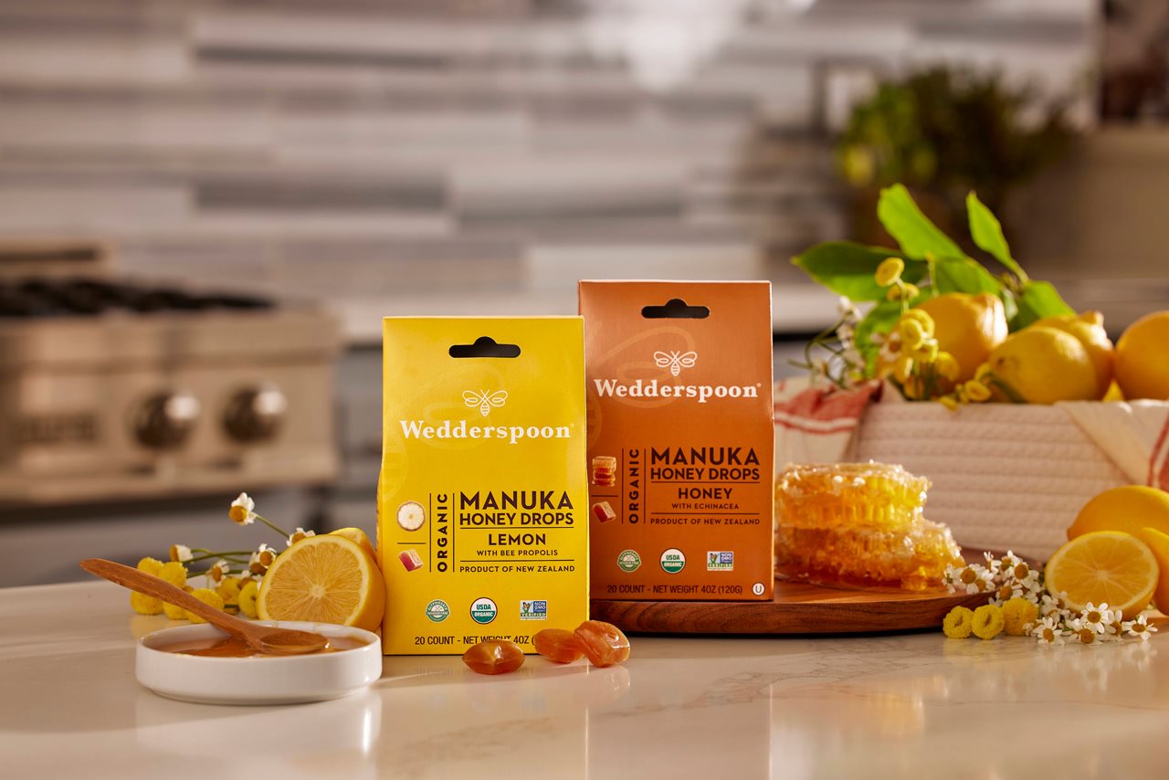 Wedderspoon  Manuka Honey and lemon and Manuka Honey drops on a kitchen counter top next to lemons and a honeycomb.