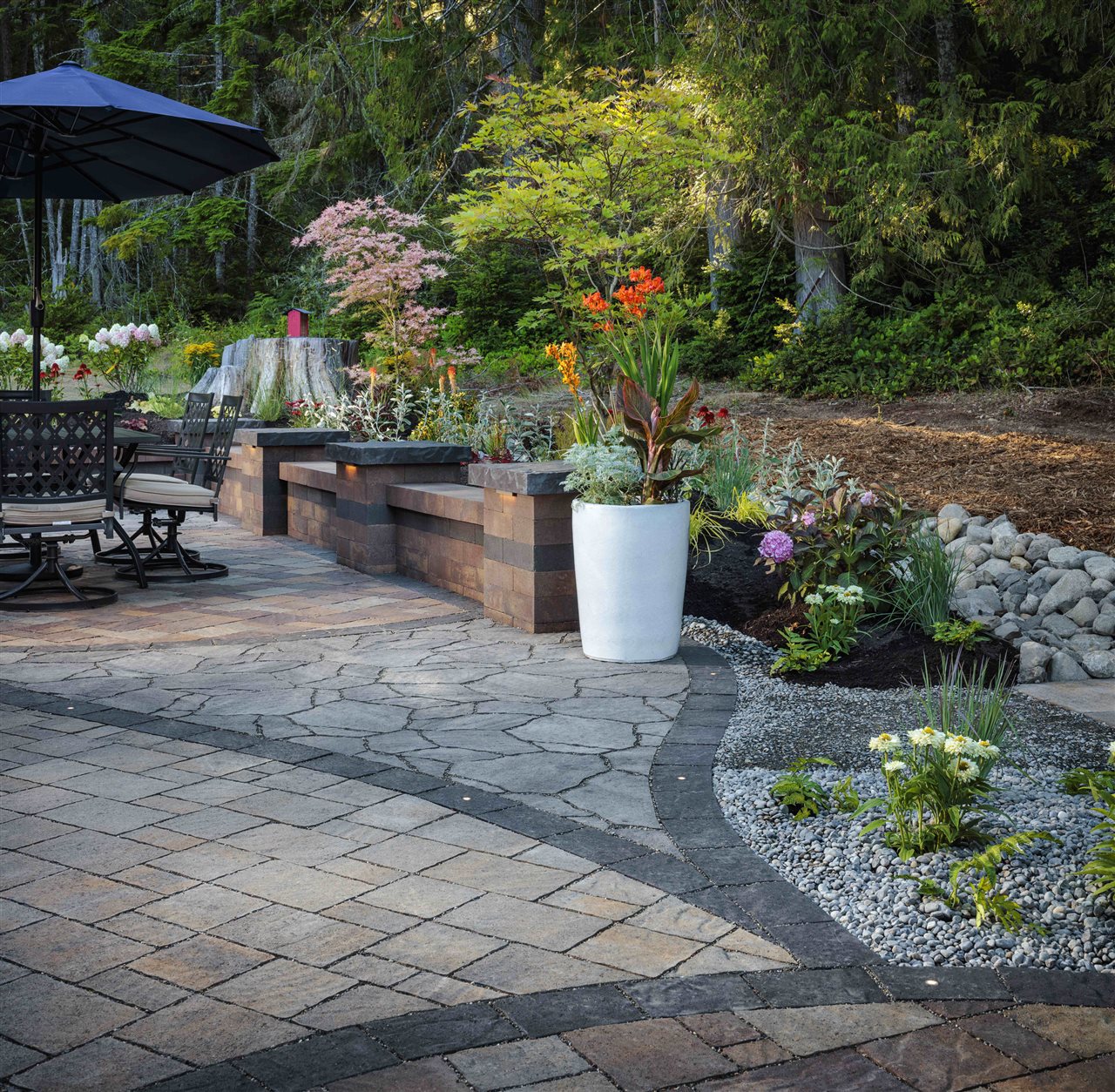 Textured paver patio with sitting wall, beautiful potted plants, and a patio table with umbrella.