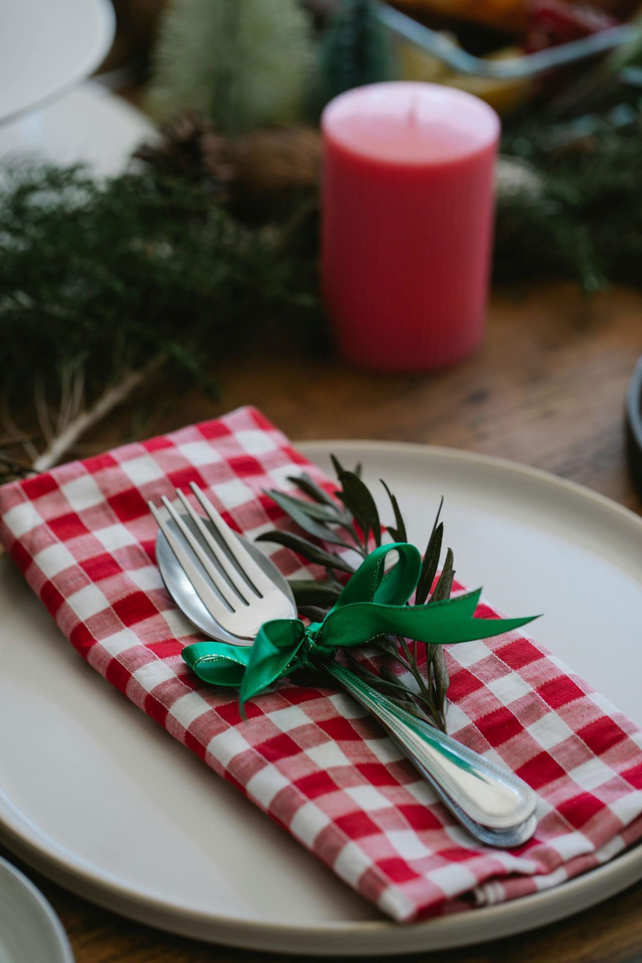 Red and white checkers napkin with flatwear wrapped in green ribbon on a plate next to natural greens on a holiday table.