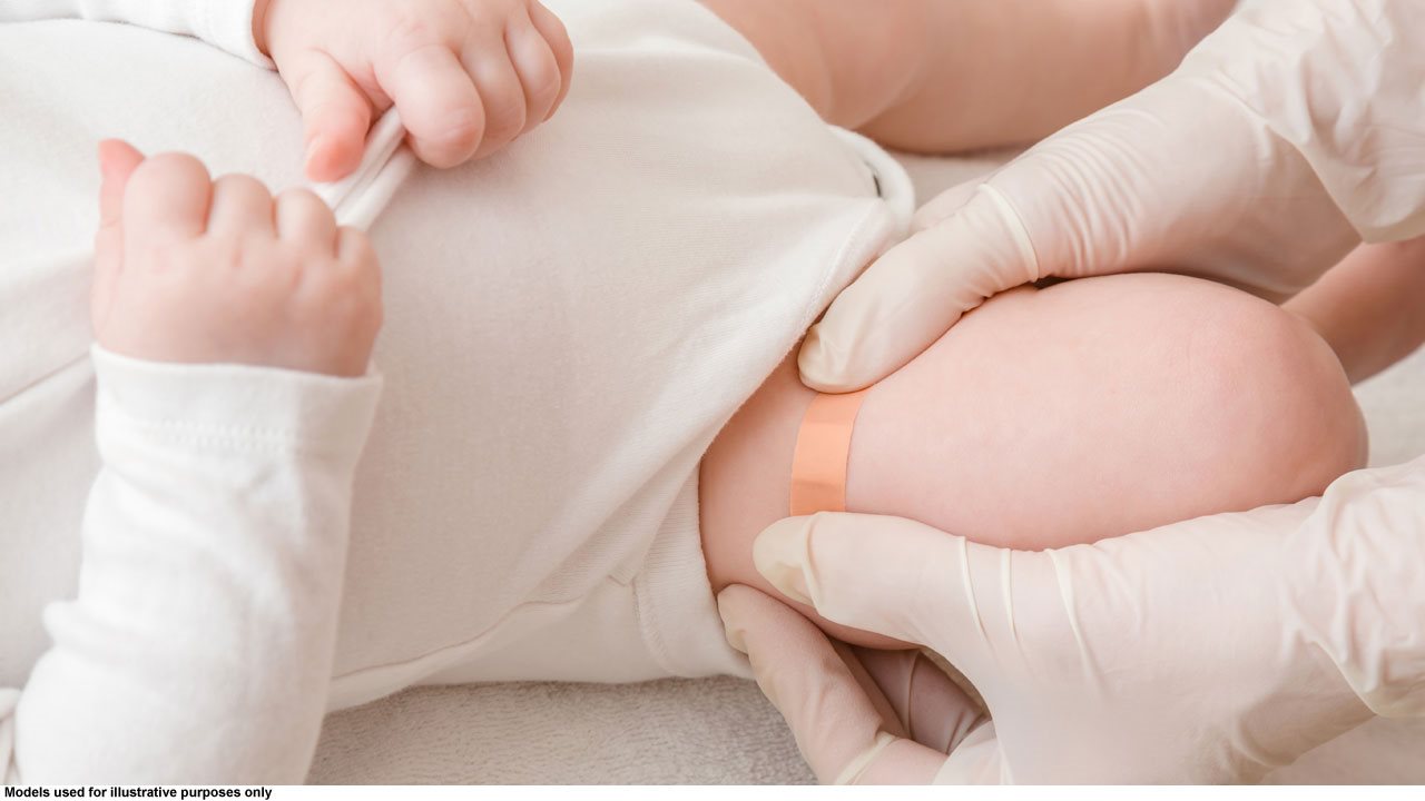 Healthcare worker wearing rubber gloves placing a bandage on a baby's leg after having given her a vaccination.