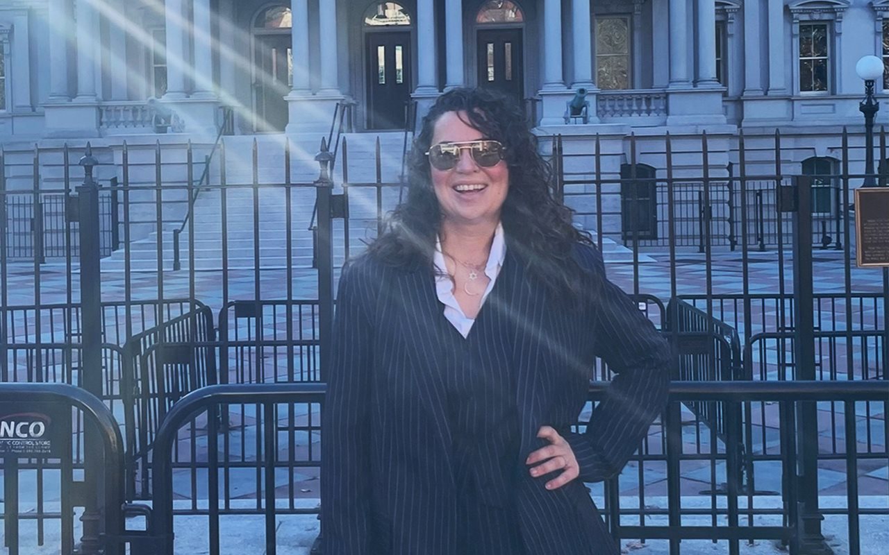 Smiling Lindsay wearing aviator sunglasses and a three piece blue pinstriped suit standing outside a beautiful building with a rod iron fence.