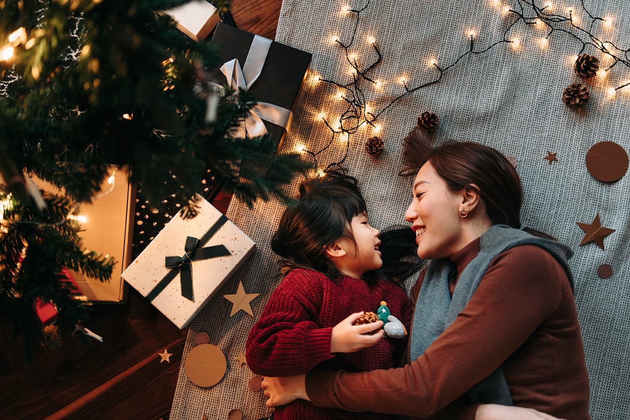 Mother and small child curl up on the floor next to the Christmas tree with lights and packages all around.