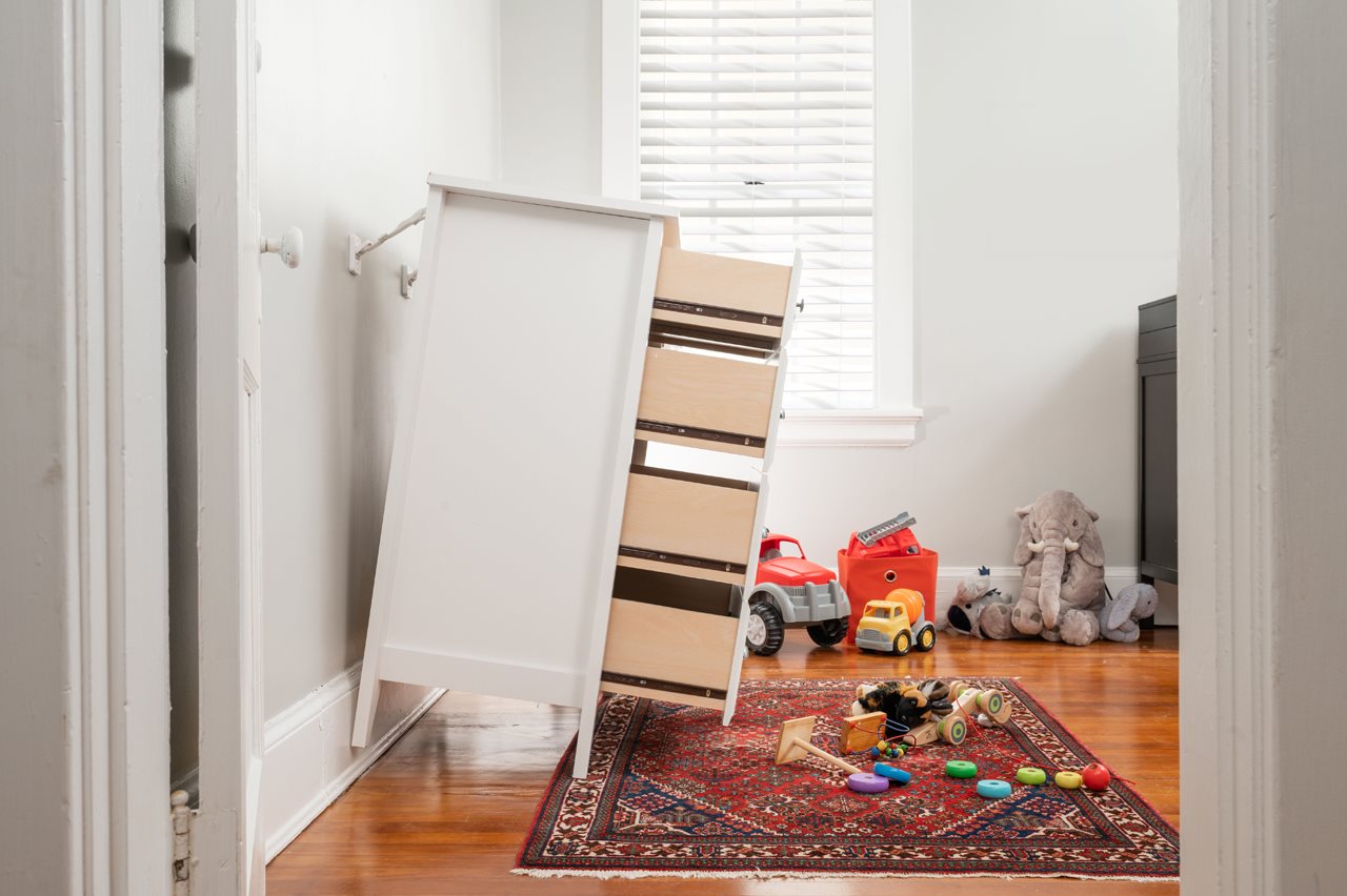 A dresser with open drawers tilted away from the wall and held in place by a device mounted to the wall that does not allow the dresser to fall.