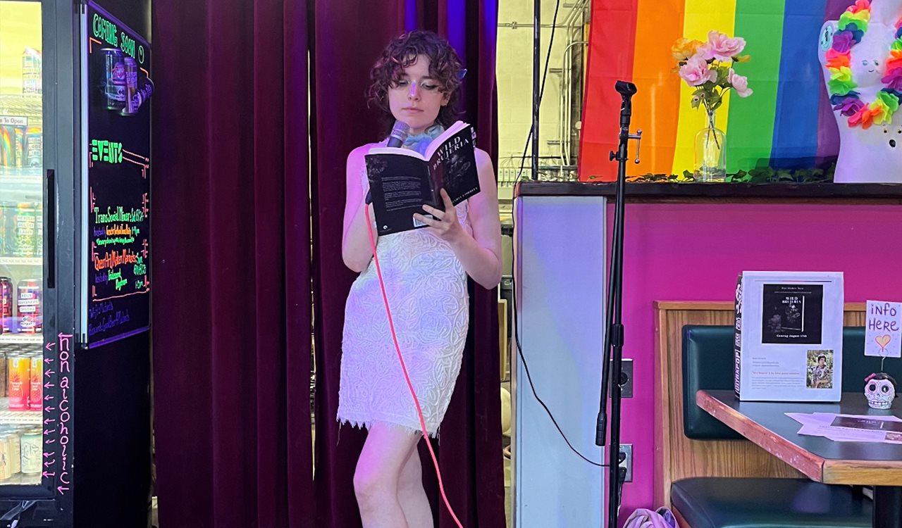 Tatiana in a white cocktail dress holding a microphone and reading her book to a group of people at a venue.