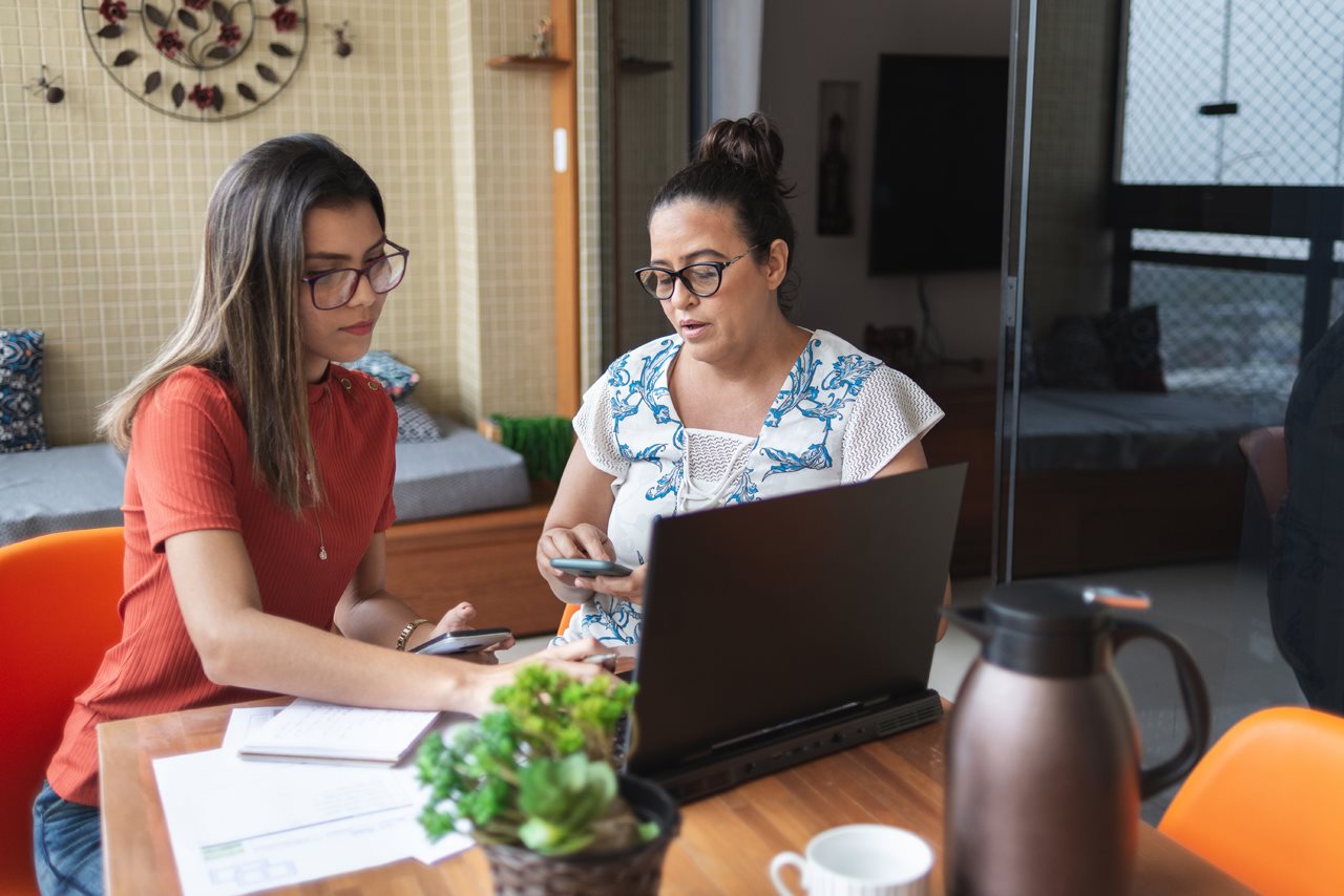 Mother and daughter both working on calculators and referencing laptop information regarding college expenses.