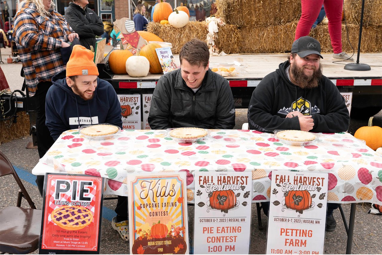 Three smiling men sit at a table preparing for a pie-eating contest.