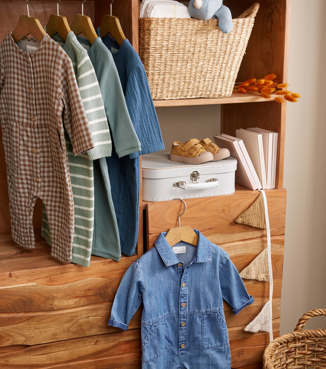 4 little outfits hanging in a closet near a basket with stuffed animals on one shelf and books and cute little shoes on the other shelf.