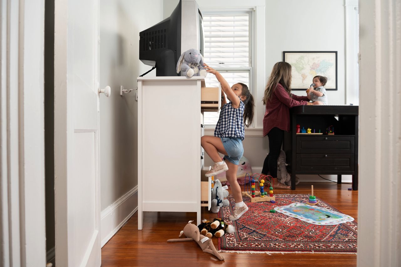 Little girl climbing up the open drawers of a dresser that has a large TV on top which is secured straps to the wall so it will not tumble over on her.