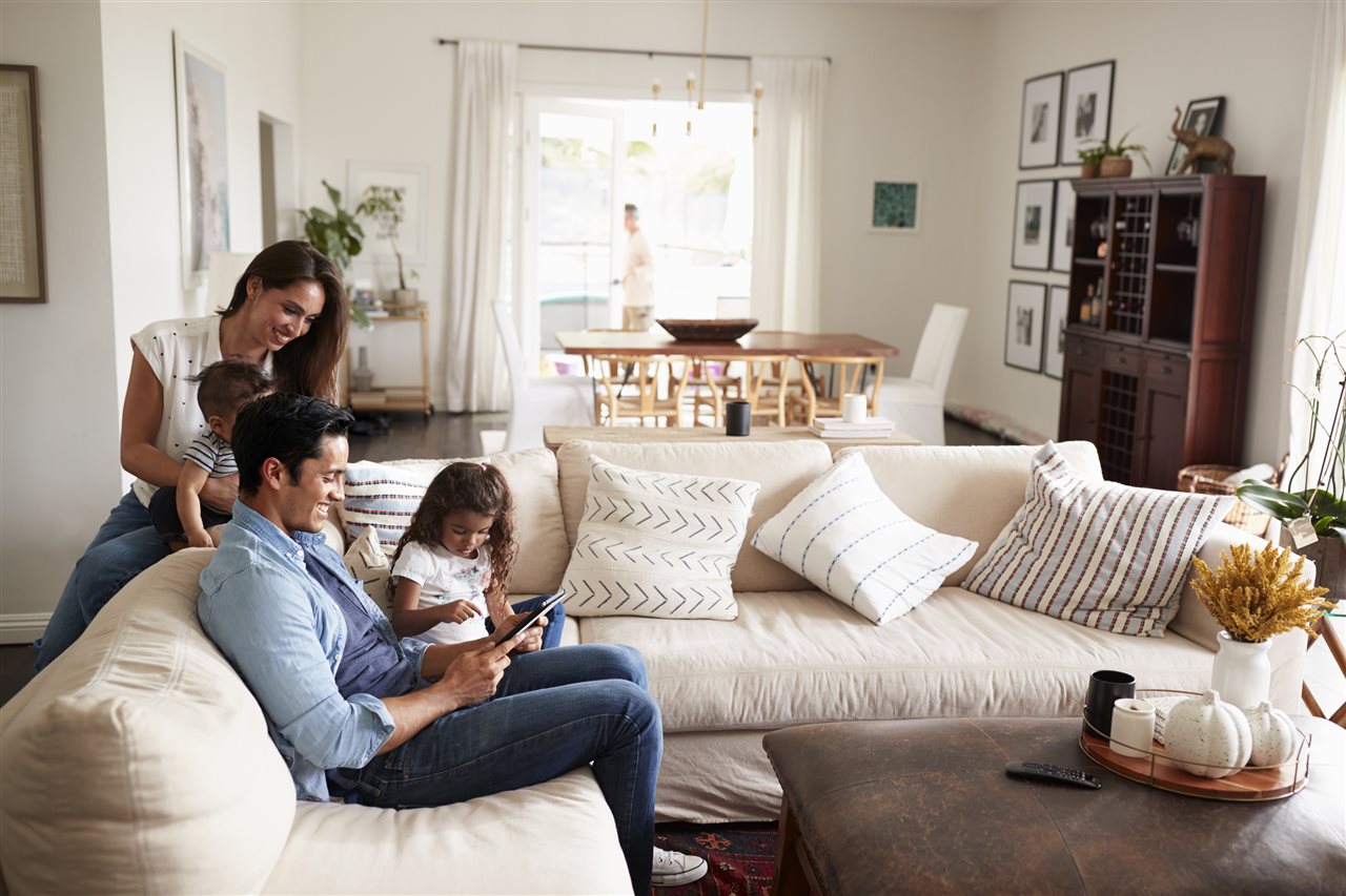 Smiling mom, dad and little girl sitting on white sectional couch in the living room using a laptop.